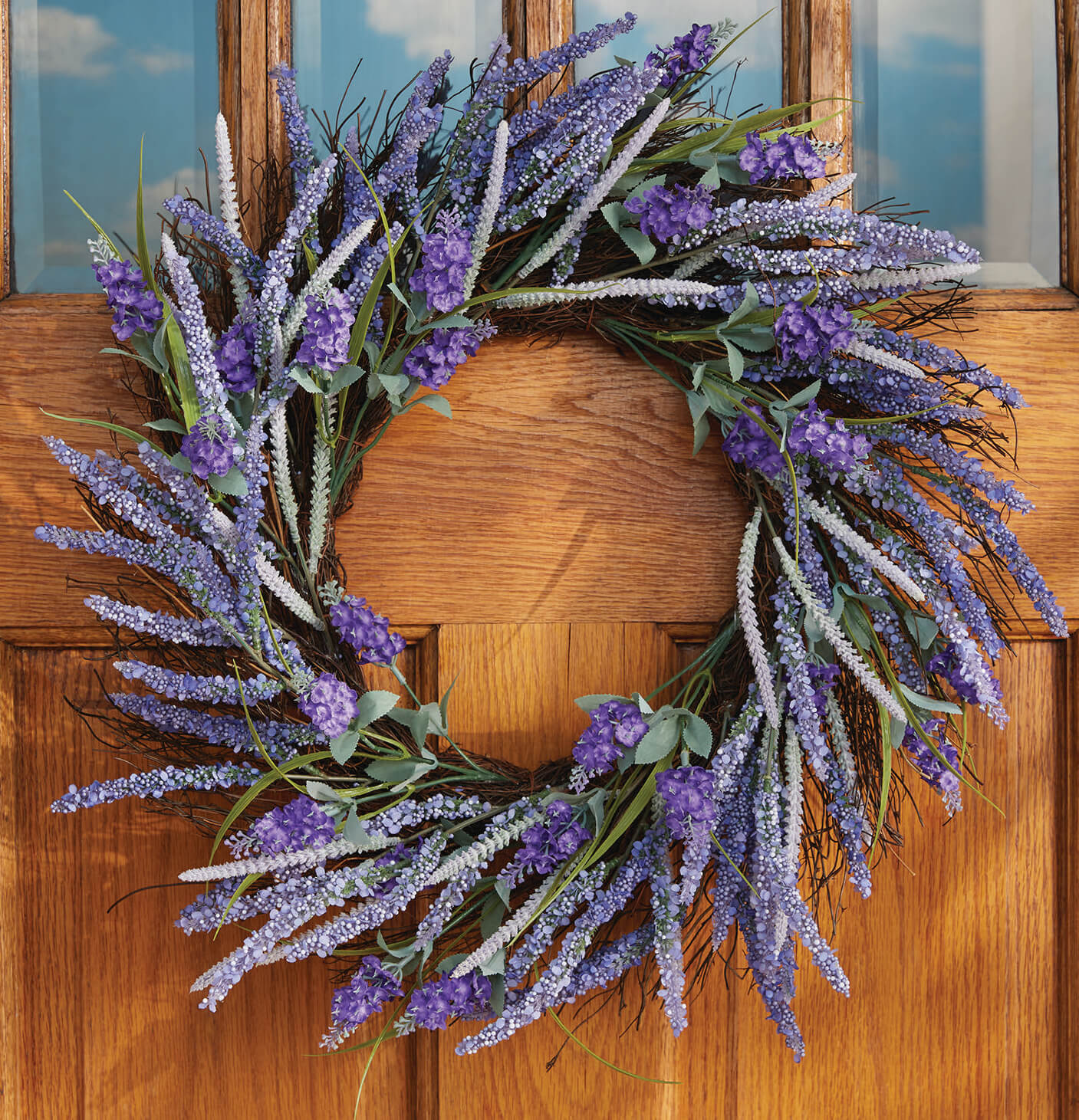 A full, round floral twig wreath of lavender hung on an oak door with paned windows.