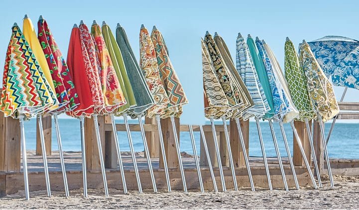 Twenty patio umbrellas in different colors and patterns leaning on an oceanside beach fence.