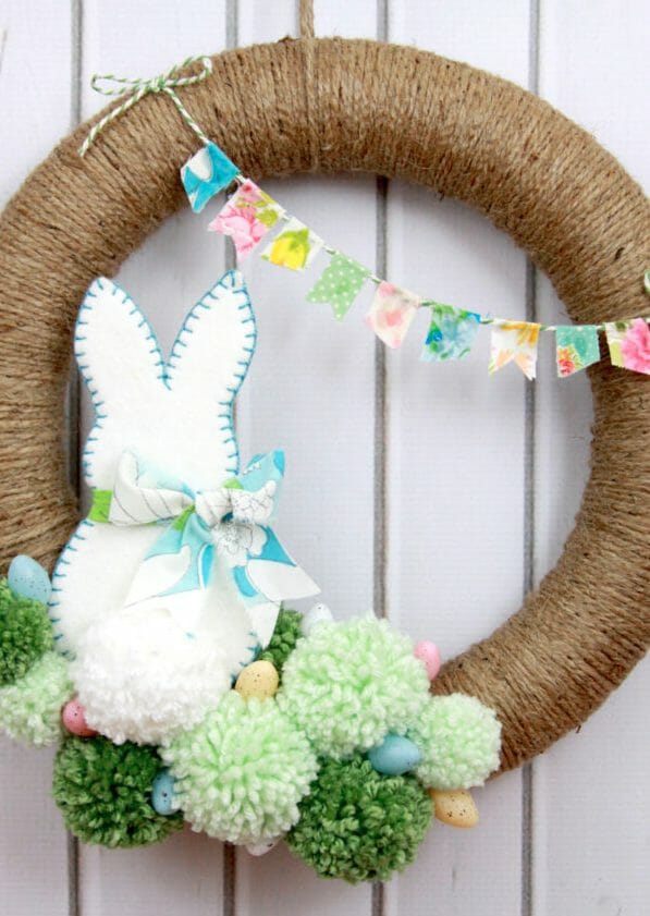 A round twine-wrapped Easter wreath with a felt bunny sitting on green pom poms, colored eggs, and a floral garland.