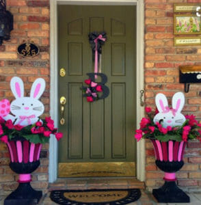 Two hot pink and magenta planters with flowers and Easter bunny cutouts by a front door, and a floral B door hanger.