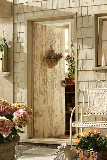 Rustic front door with a basket of flowers, a white woven metal bench, and planters of flowers.