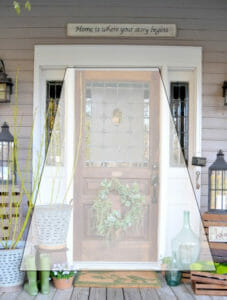 Farmhouse front porch with wreath, galvanized metal planters, wood crates, and lanterns, with a screened trapezoid overlay.