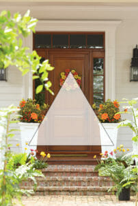A front door with an orange floral wreath, brick steps, tall planters with orange flowers, and a screened triangle overlay.