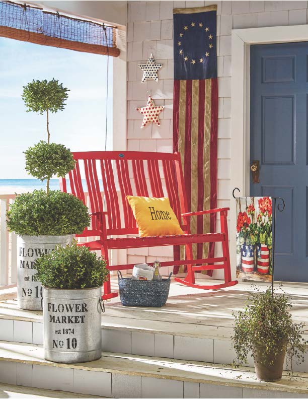 A porch with a red wooden rocking bench, galvanized buckets with boxwood, a US 13-star banner, and an ocean view.