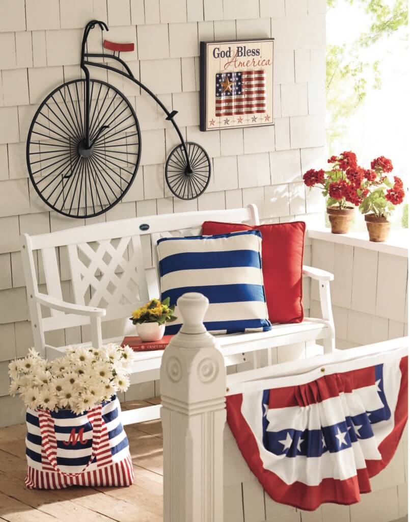 Porch with a white bench, red and blue stripe pillows, patriotic banner, red geraniums, vintage wall bike.