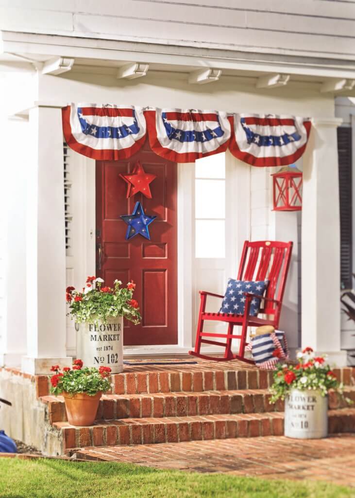 A red front door with stars, Patriotic bunting, a red rocker with pillows, brick steps and three red geranium planters.