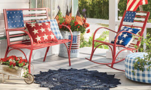 Red metal USA flag bench and rocker with red and blue star pillows, and a galvanized can of red geraniums and five USA flags.