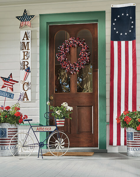 Patriotic front porch with an America sign, a bike planter with white geraniums, a tall US 13-star banner, and red geraniums.