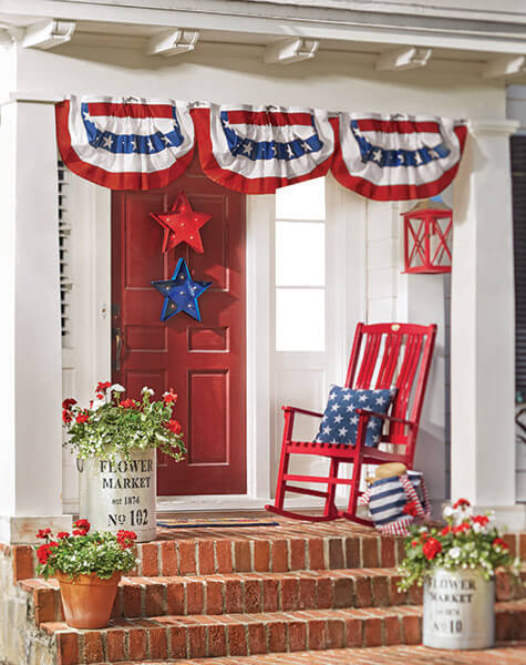 Patriotic front porch bunting, red and blue marquee lights on the red door, a red rocker with a pillow, and red geraniums.
