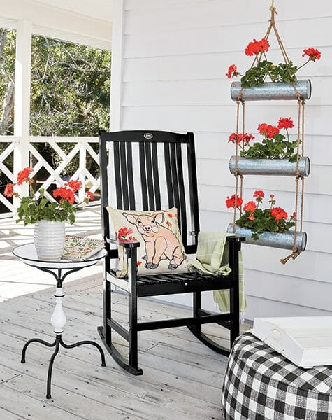 Farmhouse porch with black rocker, pig pillow, black gingham ottoman, and red geraniums in a hung 3-tier metal planter.