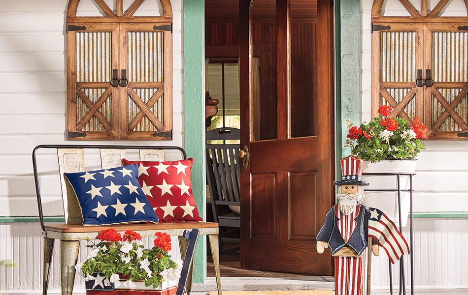 Patriotic front porch with red and blue star pillows on a metal bench, an Uncle Sam wood cutout, and red geraniums.