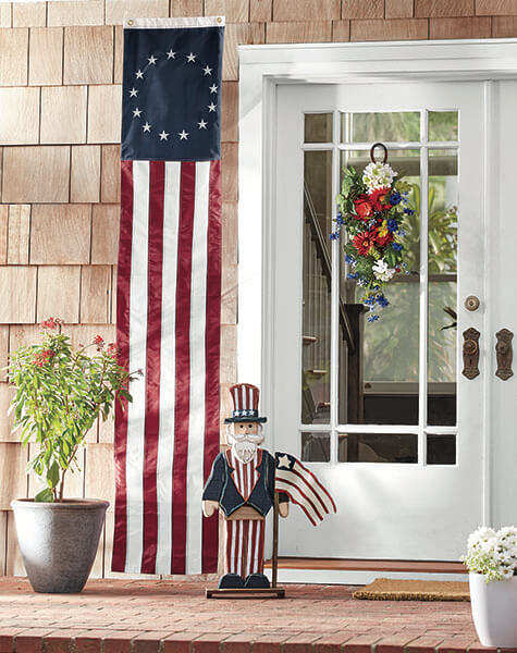 A patriotic porch with a red, white, and blue swag on a white door, a US 13-star banner, and a wooden Uncle Sam.