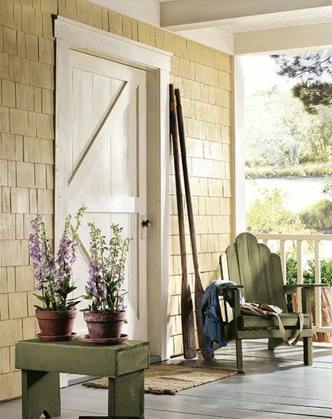 A Farmhouse porch with yellow cedar shakes, a green adirondack chair, two oars, and a green bench with potted delphiniums.