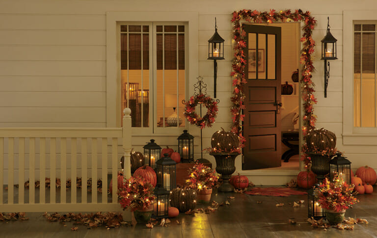 A front door with lit Fall leaves garlands, three planters, and a wreath, with eight lit black lanterns and several pumpkins.