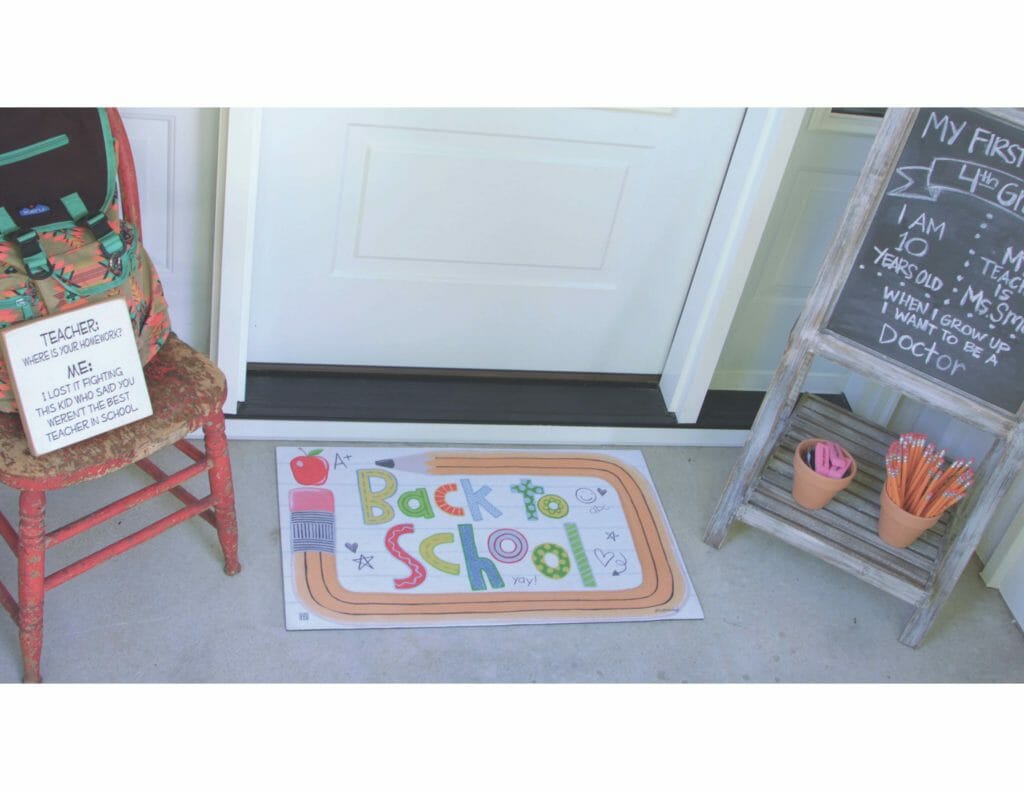 A back to school door mat, a wooden distressed chalkboard easel, and a red distressed chair with a versed Teacher sign.