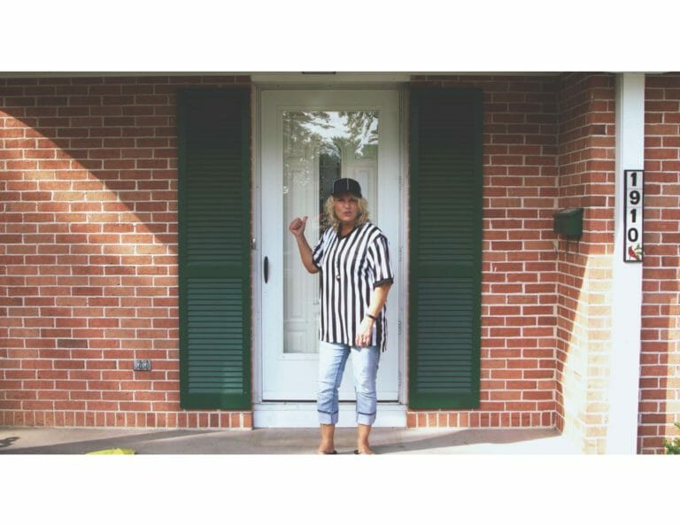 A blonde woman standing by a front door, dressed in a black and white referee shirt and a black hat.