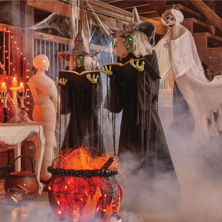 Life sized witches with shiny apples in hand stand next to a hanging white ghost skeleton and other Halloween decor