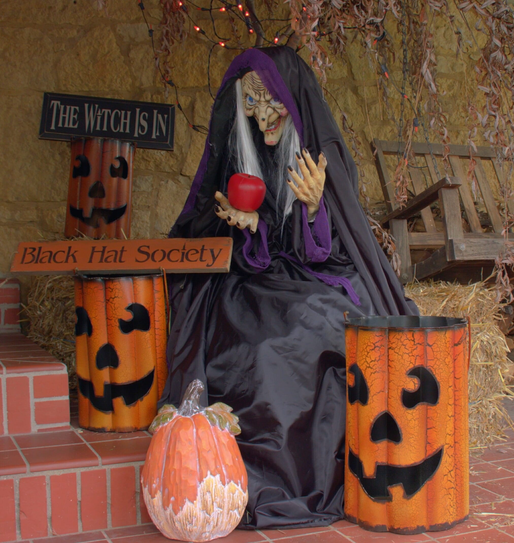 A witch sitting on straw bales with apple in hand, next to three metal Jack-o-Lantern luminaries and Halloween signs.