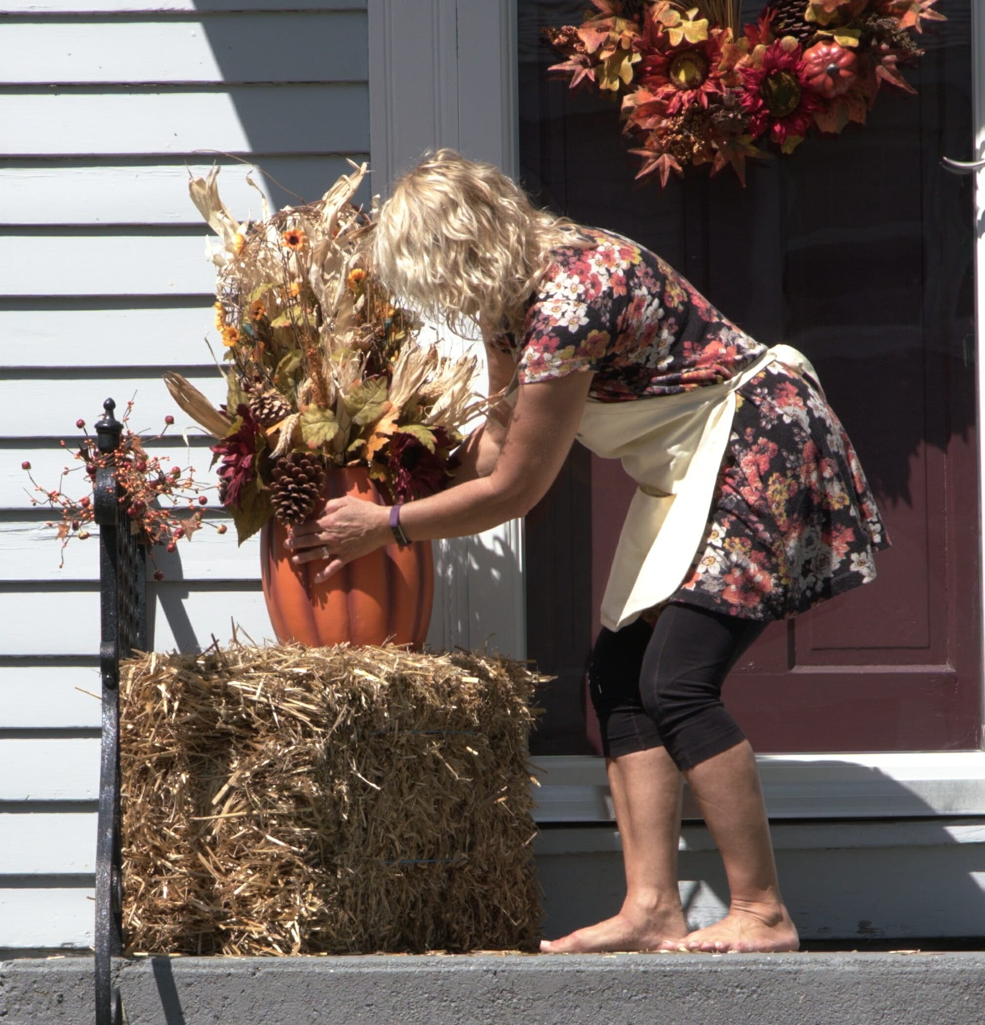 woman arranging a pumpkin planter full of corn husks pine cones and other fall décor on top of a bale of straw