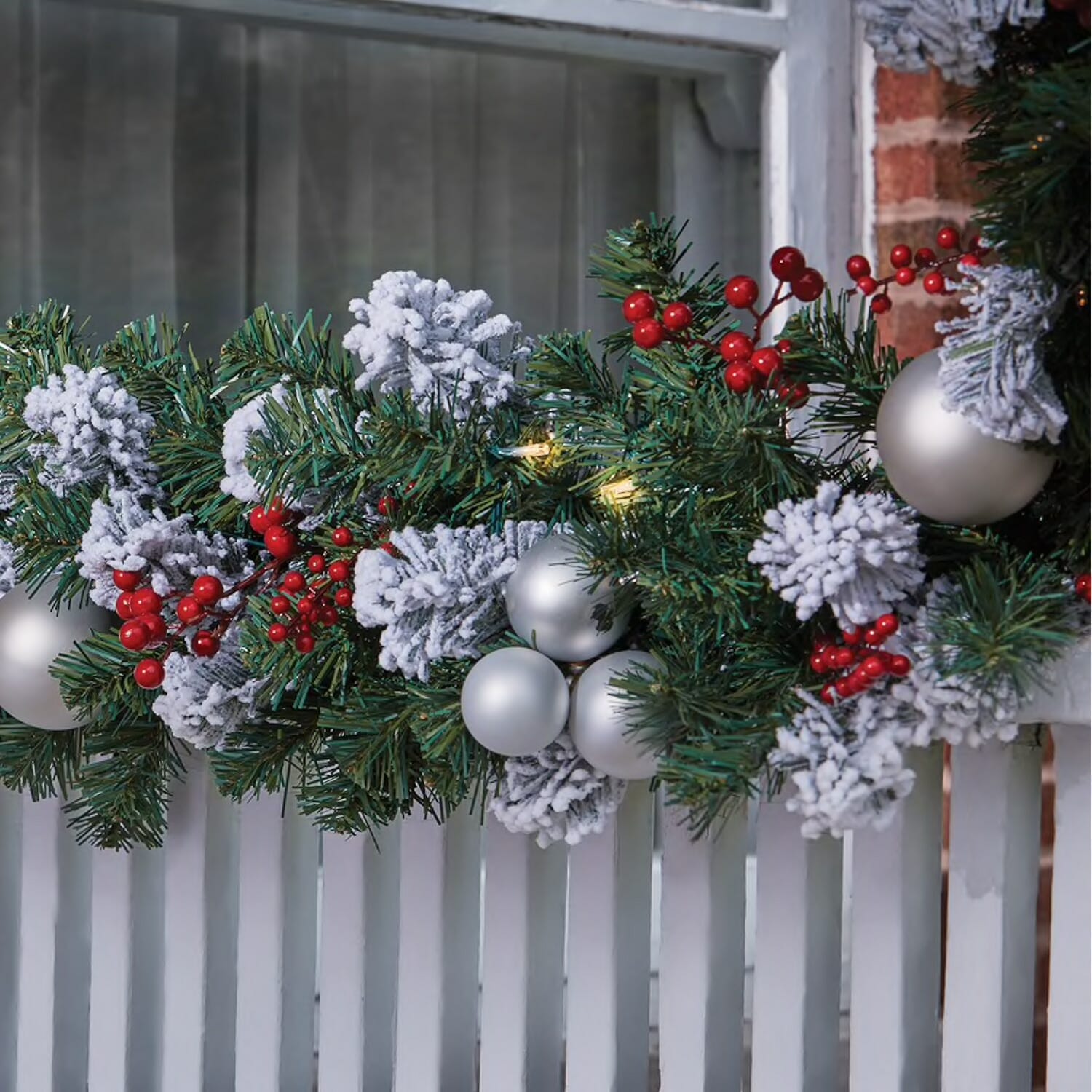 A lit garland on a porch railing, with flocked and green pine, white ball ornaments, and red berries.