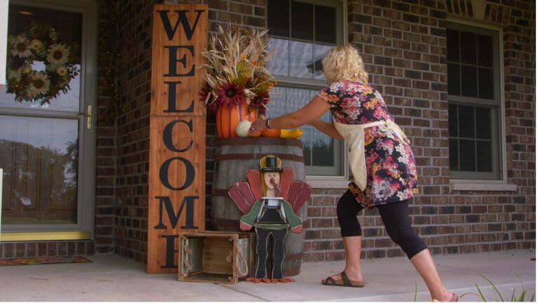 woman placing pumpkins and gourds on a wooden barrel next to vertical welcome sign and fall décor on porch