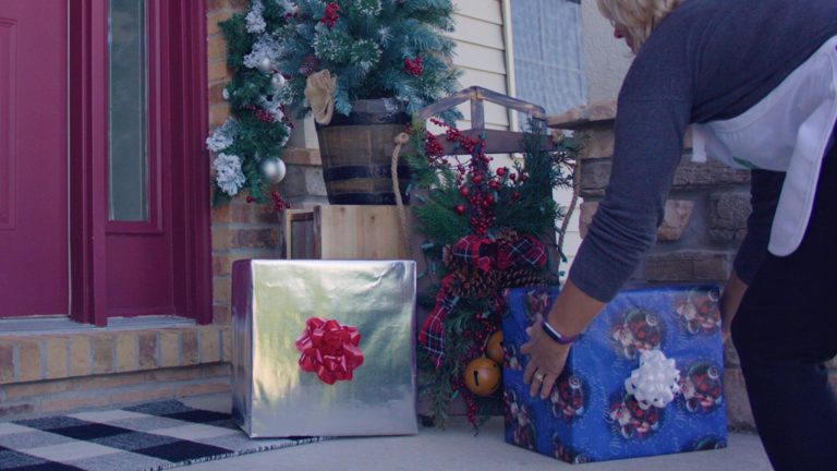 woman putting presents by wooden sleigh with red plaid ribbon and evergreens to create a Country Christmas porch