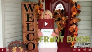 woman on porch with vertical welcome sign, orange red yellow garland, wreath with gourds and other fall décor