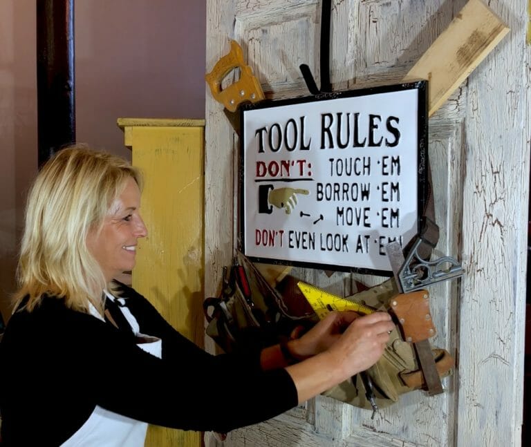 woman hanging a wreath that consists of a tool rules sign with toolbelt and other tool décor