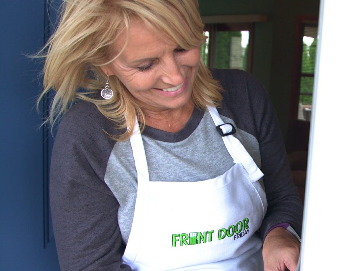 A smiling blonde woman in a white Front Door apron, applying weather stripping to a white door jamb.