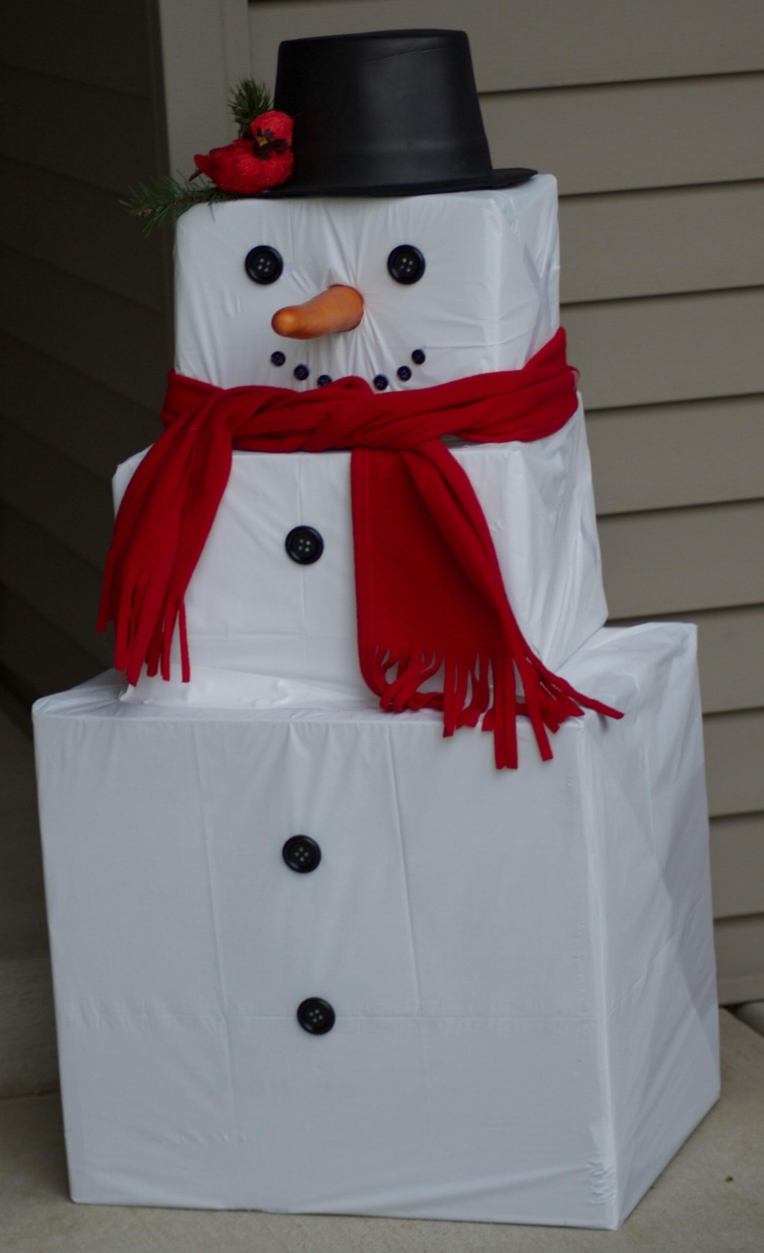 Three square boxes sitting on porch create Snowman with red scarf, carrot and cardinal sitting on hat