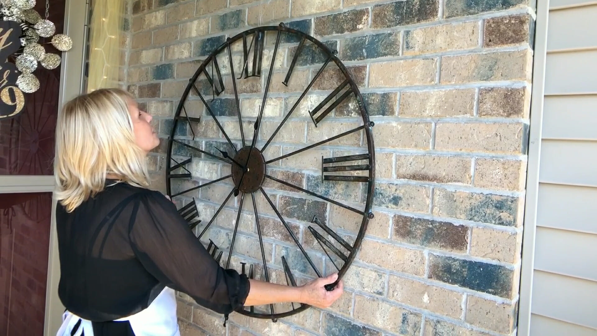 woman hanging a very large metal clock on front porch