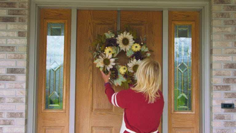 A blonde woman placing a round Fall wreath of beige sunflowers, pine cones and yellow gourds on an oak front door.