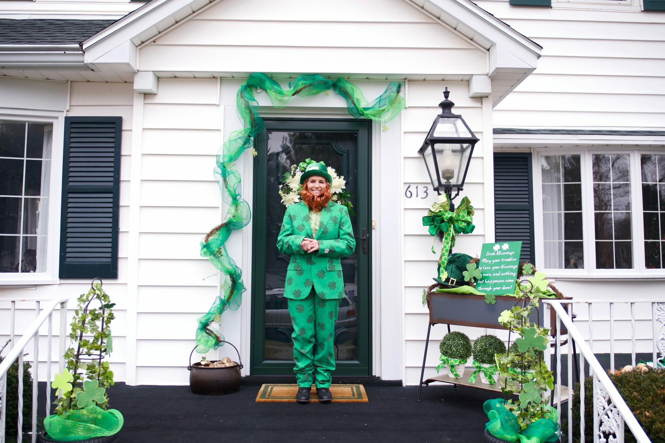 woman dressed as leprechaun in front of house with pot of gold Irish blessing sign green topiaries St. Patrick's Day décor