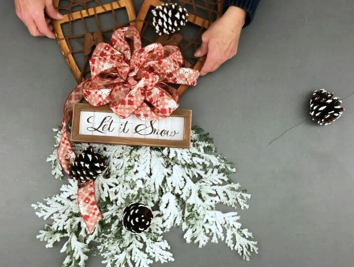 woman adds pinecones to winter wreath consisting of flocked evergreens red and white ribbon and wooden snowshoes