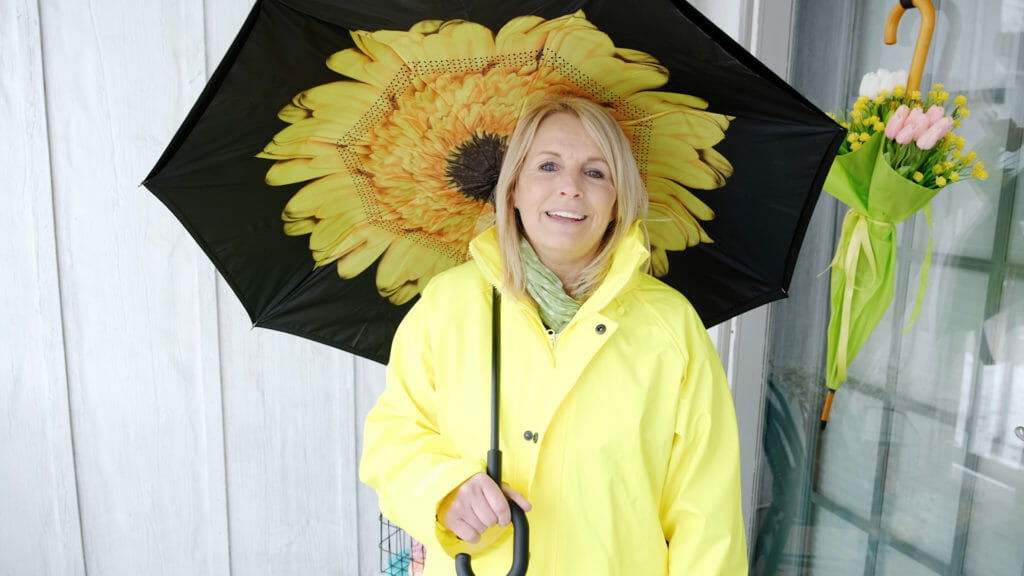 A smiling woman in a yellow raincoat under a yellow flower umbrella, next to a closed umbrella door swag holding tulips.