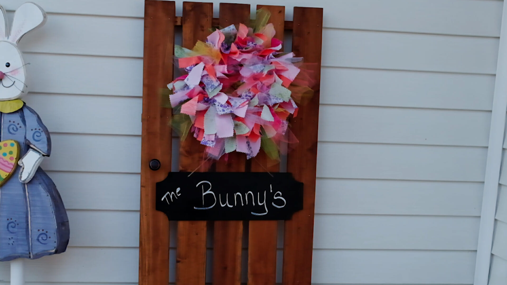 A wood fence section leaning against house siding, with a colorful rag wreath and a sign for The Bunny's.