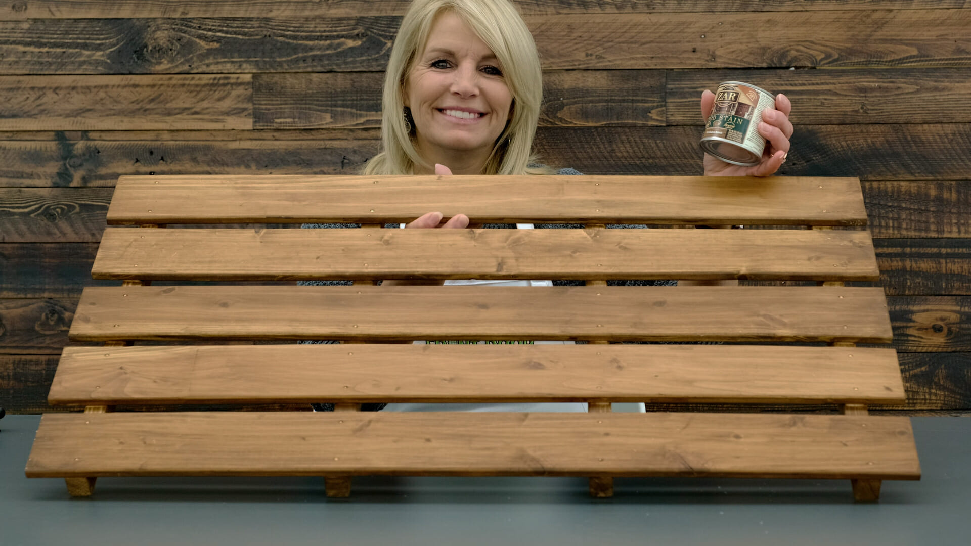 A smiling woman holding a section of a short wood picket fence and a can of wood stain.