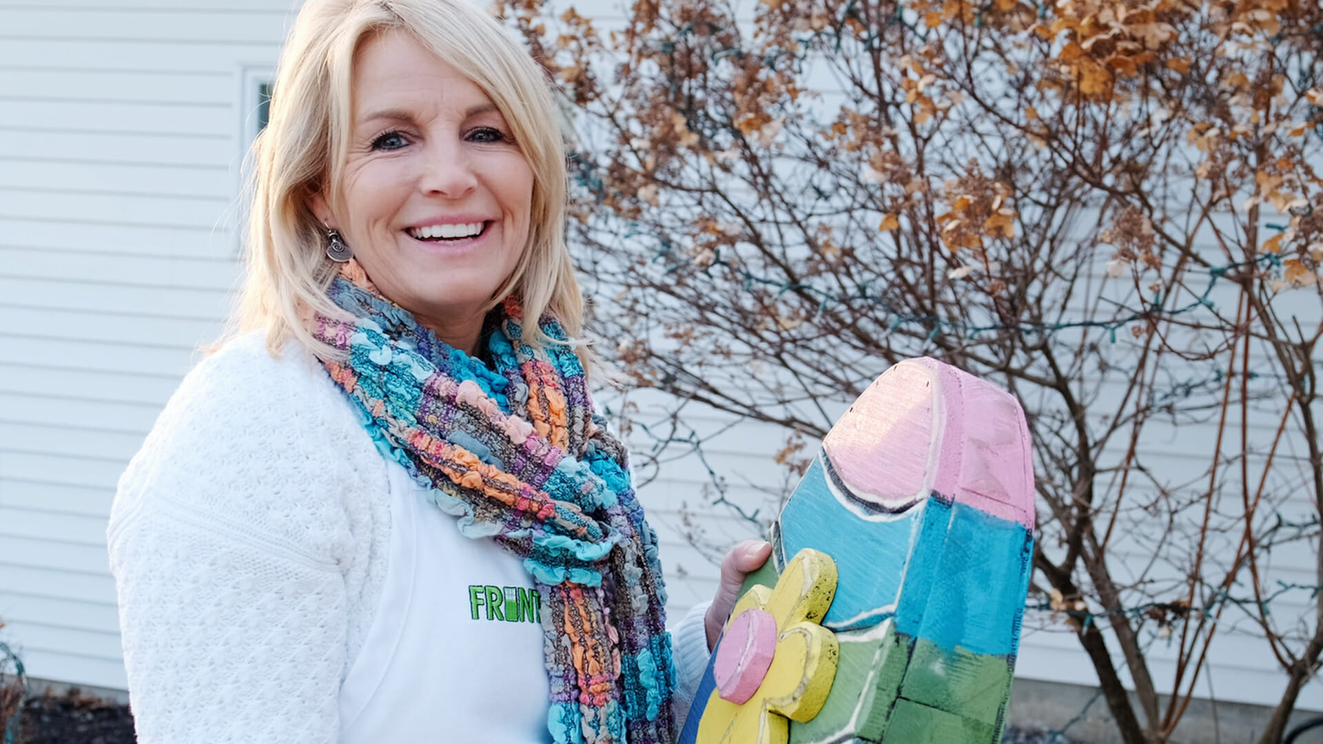 A smiling woman holding a large colorful wood Easter egg with a wood flower on the front.