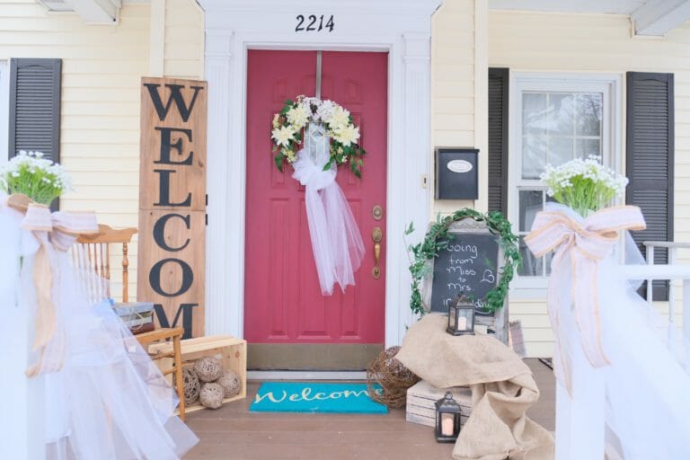 White floral wreath on a red door, a tall WELCOME sign, potted white daisies with long white tulle bows, and a chalkboard.