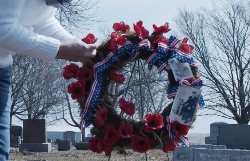 Placing a poppy wreath with photos and patriotic ribbon at a cemetery gravesite.