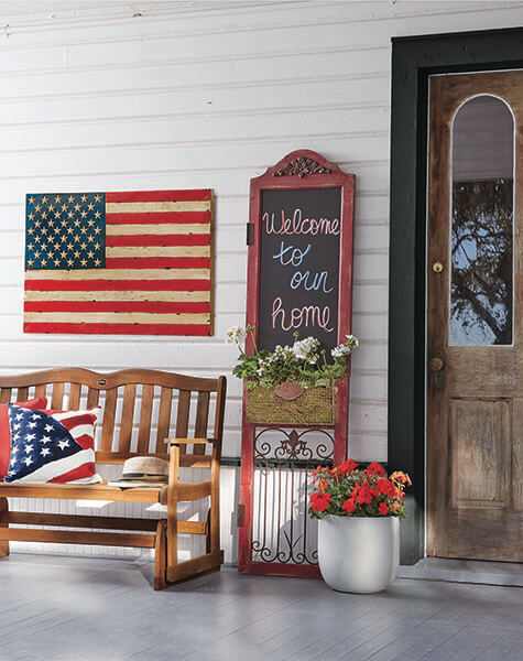 Farmhouse front porch with a wood bench and flag pillow, a large USA flag plaque, a chalkboard shutter, and red geraniums.