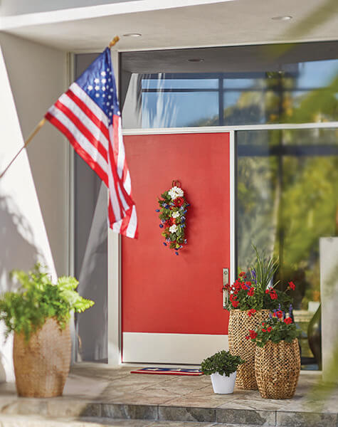 A modern red door with a red, white and blue swag, tall woven planters with red geraniums, a fern, and a US flag.