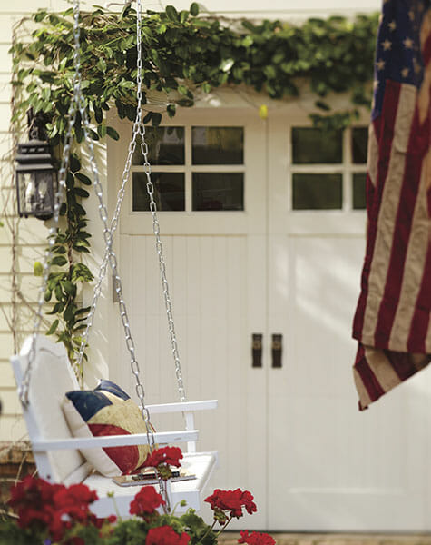 Ivy over front double Farmhouse doors, with a hanging white swing, star pillow, flag, and blooming red geraniums.
