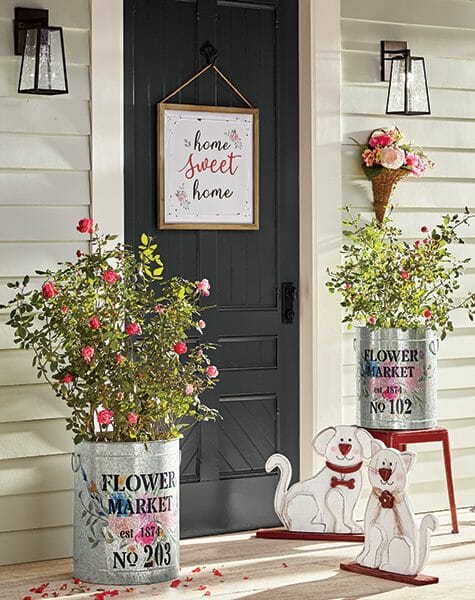 Farmhouse front door with Home Sweet Home plaque, market tins of blooming roses, floral hung basket, two wood pet cutouts.