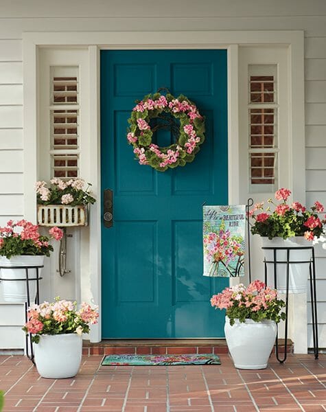 A teal front door with a pink geranium wreath, five planters of pink geraniums, a floral teal door mat & matching flag.