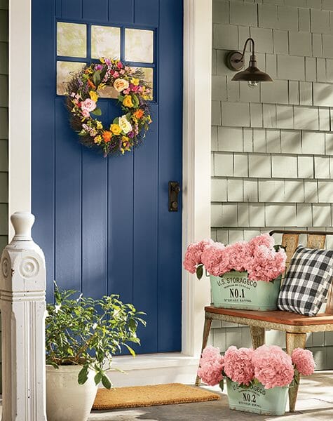 A navy front door with a round floral wreath, two seafoam metal pails filled with pink hydrangeas, and a wash tub bench.