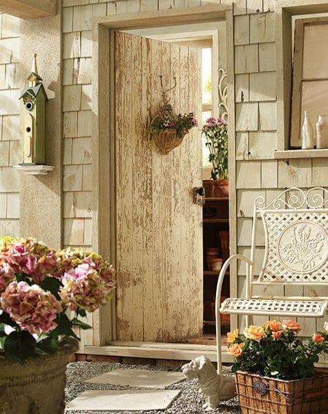 A rustic front door with a basket of flowers, a white woven metal chair, a tall church birdhouse, and planters of flowers.
