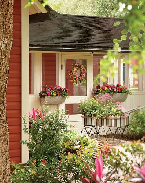A red cottage with white trim, a wreath and two matching window boxes of red flowers, a cart and walk with mixed flowers. 