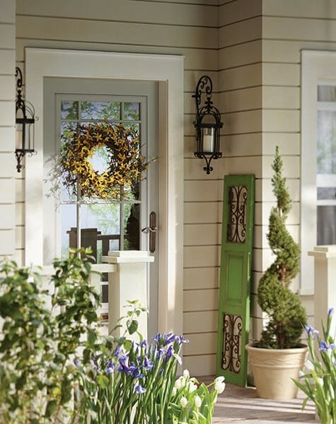 A Springtime front porch with a forsythia wreath, a scrolled shutter, hung lanterns, and blooming iris and white tulips.