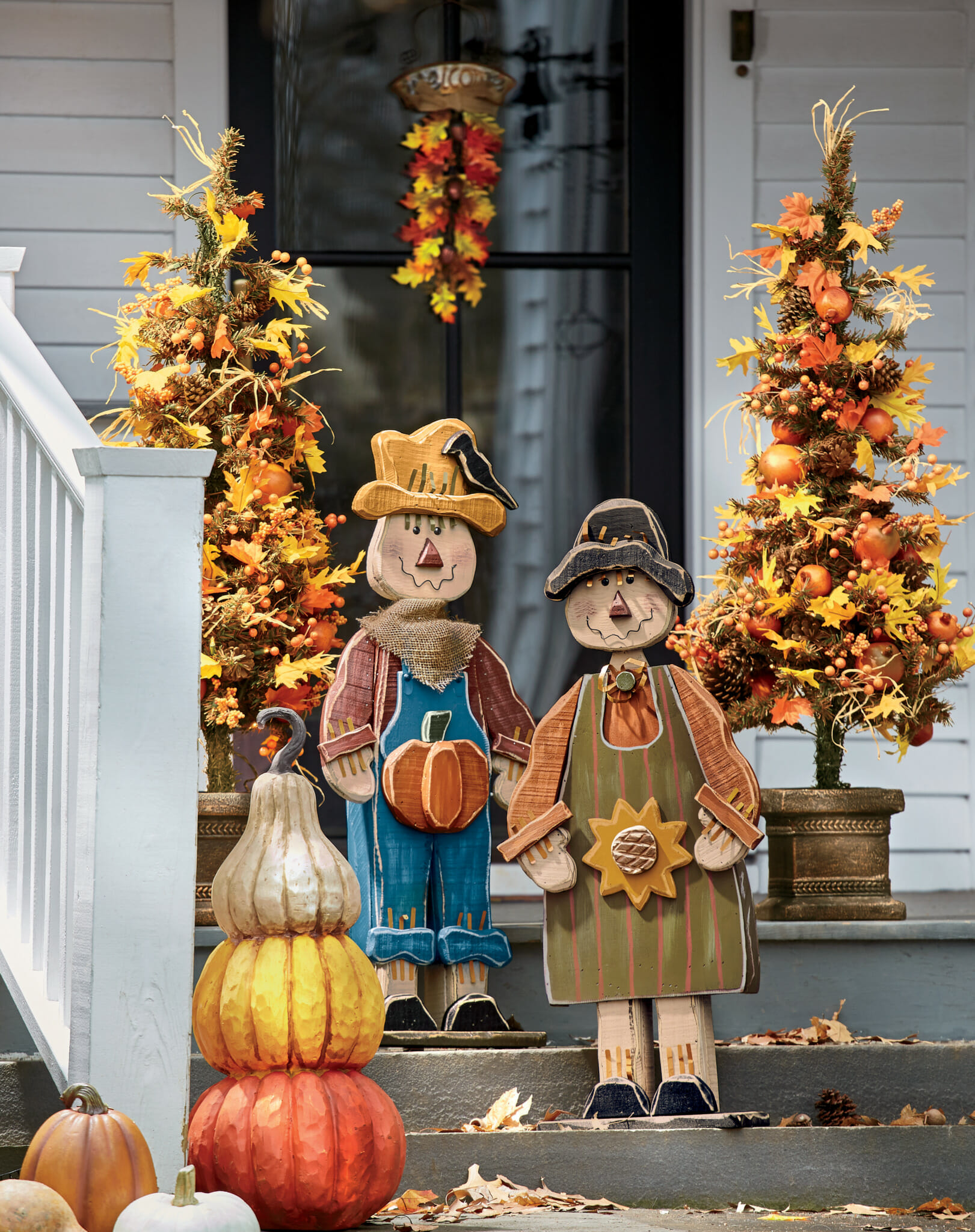 Wood cutout scarecrow couple on a front porch with stacked pumpkins and Fall leaves trees and swag.